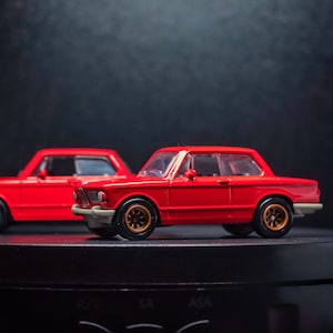 May include: Two red toy cars, one behind the other, on a black surface. The car in the foreground is a classic coupe with a black interior. The car in the background is a saloon.