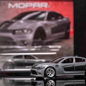 May include: A grey Dodge Charger diecast model car with black racing stripes and the word "MOPAR" on the side. The car is sitting on a reflective surface.
