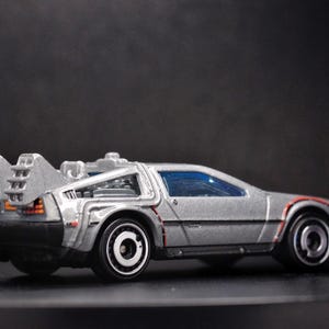 May include: A silver toy car with a futuristic design, featuring a rear-mounted engine and a large spoiler. The car has a blue tinted windscreen and black wheels.