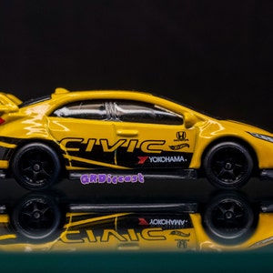 May include: Yellow Honda Civic Type R diecast car with black and white racing stripes, "CIVIC" and "YOKOHAMA" logos, and a Hot Wheels logo. The car is on a reflective surface.