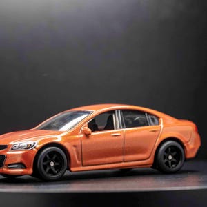 May include: A bright orange toy car model of a sedan with black wheels. The car has a black interior and a black grill.