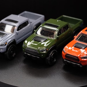 May include: Three toy pickup trucks in different colors: gray, green, and orange. The trucks are all facing forward and have black wheels.