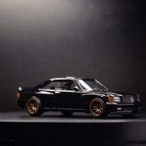 May include: A black toy car model with gold wheels. The car is a coupe with a sleek design and is photographed on a black surface.
