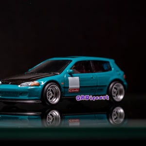May include: A teal blue Honda Civic diecast car model with a black hood and white racing stripe. The car has black wheels with silver rims and the word "Civic" is printed on the side. The car is sitting on a reflective surface.