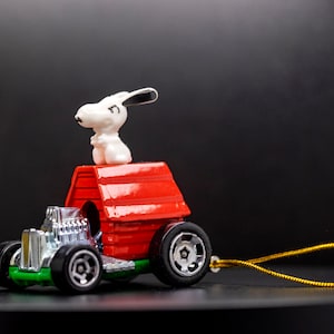 Op de afbeelding: Een rode speelgoedauto met een wit hondenfiguurtje erop. De auto heeft een zilveren grille en zwarte wielen. De hond zit op een rood hondenhok dat aan de achterkant van de auto is bevestigd. De auto heeft een gouden touwtje eraan.