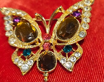 Puede incluir: Un broche de mariposa de tono dorado con diamantes de imitación transparentes, marrones, rosas, morados y azul verdoso.