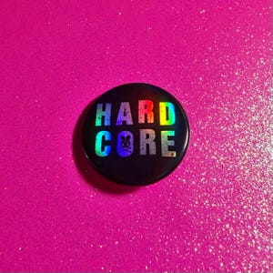 May include: A black, round button with the words "HARD CORE" in a rainbow holographic font. A small skull and crossbones graphic is below the word "CORE". The button is set against a bright pink, glittery background.
