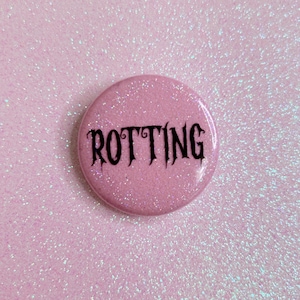 May include: A round, light pink button with the word "ROTTING" in black gothic font. The button has a glittery finish and is set against a matching glittery pink background.