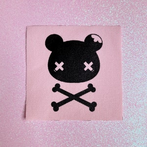 May include: A square, light pink fabric patch featuring a black cartoon bear head with crossbones below. The bear has crossed-out eyes. The patch is likely for sewing or ironing onto clothing or accessories.