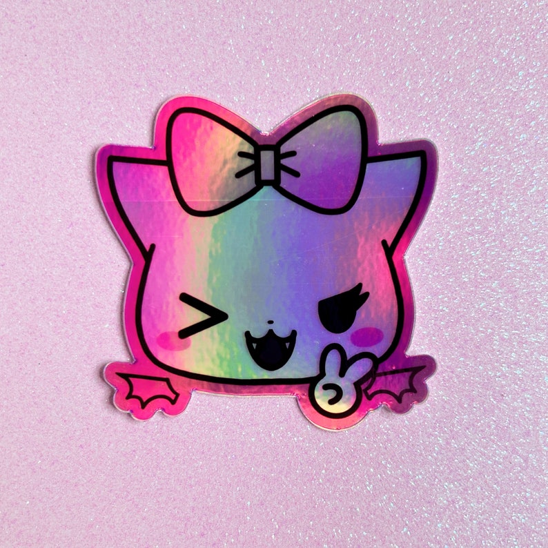 May include: A holographic sticker of a cartoon cat with a rainbow gradient, wearing a bow and winking. The cat has a peace sign and bat wings.