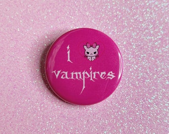 May include: A pink button with a white outline and the text "I love vampires" with a cartoon vampire kitty in a heart shape above the text.