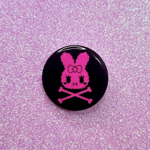 May include: A black button with a pink glitter bunny wearing a bow and crossed bones. The bunny has a cute expression.