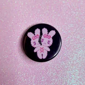 May include: A pink and white stitched together bunny with black eyes and a black background. The bunny has a stitched seam down the middle of its body.