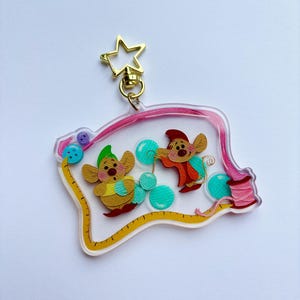 Könnte beinhalten: Ein klarer Acryl-Schlüsselanhänger mit zwei Zeichentrickmäusen und Näh-Elementen. Der Schlüsselanhänger hat einen goldenen Sternverschluss und einen rosa Bandrand. Die Mäuse sind von türkisfarbenen Blasen, Knöpfen und einem gelben Maßband umgeben.