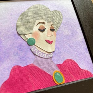 Puede incluir: Retrato enmarcado en papel de una mujer con cabello gris, vestida con un vestido rosa con un broche y aretes verdes. El fondo es un lavado de acuarela p&uacute;rpura. La obra de arte est&aacute; en un marco negro.