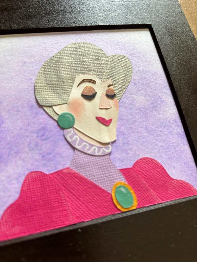 Puede incluir: Retrato enmarcado en papel de una mujer con cabello gris, vestida con un vestido rosa con un broche y aretes verdes. El fondo es un lavado de acuarela p&uacute;rpura. La obra de arte est&aacute; en un marco negro.