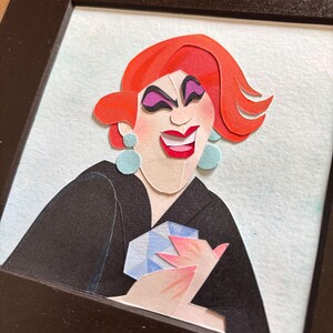 May include: A framed paper art portrait of a person with red hair, purple eye makeup, and a red smile. The person is wearing a black blazer and holding a blue and white gem. The background is light blue.