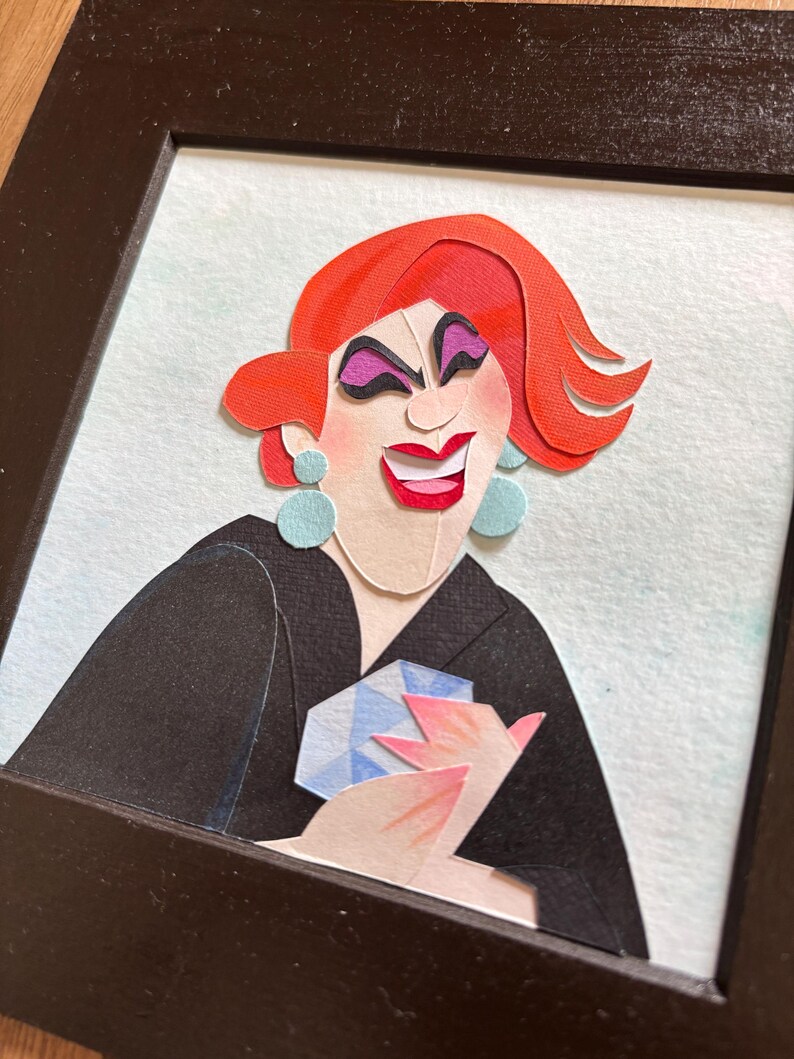 May include: A framed paper art portrait of a person with red hair, purple eye makeup, and a red smile. The person is wearing a black blazer and holding a blue and white gem. The background is light blue.
