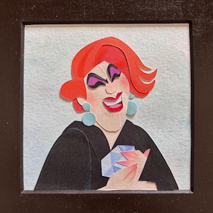 May include: Paper art portrait of a smiling person with red hair, purple eye shadow, and blue earrings. The person is wearing a black top and holding a blue object. The artwork is framed in a black square frame.