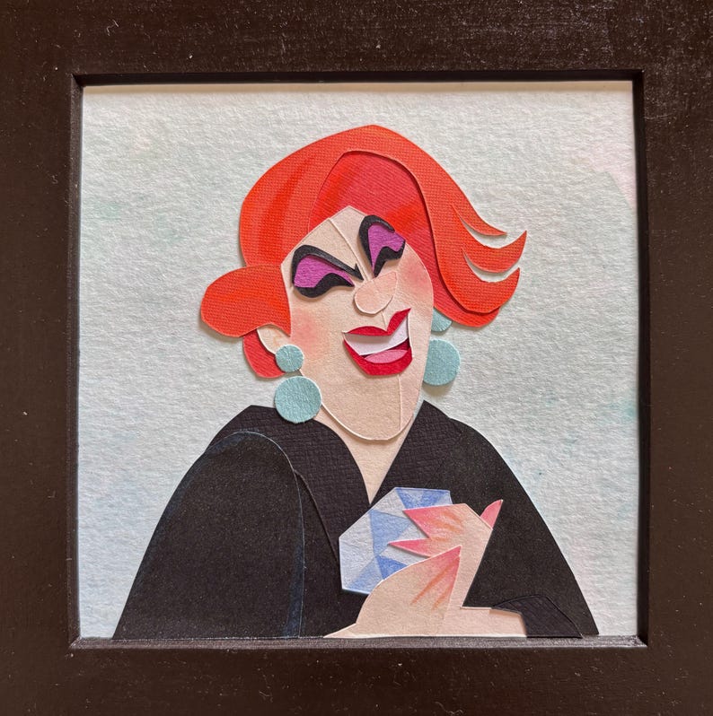 May include: Paper art portrait of a smiling person with red hair, purple eye shadow, and blue earrings. The person is wearing a black top and holding a blue object. The artwork is framed in a black square frame.