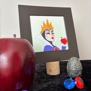 Puede incluir: Ilustraci&oacute;n enmarcada de un personaje con una corona dorada, sosteniendo una manzana roja. Una gran manzana roja, gemas decorativas y un recipiente plateado en forma de huevo est&aacute;n en primer plano. La ilustraci&oacute;n est&aacute; sobre un soporte de madera.