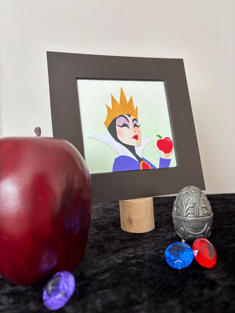 Puede incluir: Ilustraci&oacute;n enmarcada de un personaje con una corona dorada, sosteniendo una manzana roja. Una gran manzana roja, gemas decorativas y un recipiente plateado en forma de huevo est&aacute;n en primer plano. La ilustraci&oacute;n est&aacute; sobre un soporte de madera.
