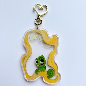 May include: A clear acrylic keyring featuring a green chameleon character with a smiling expression. The keyring has a gold-coloured heart-shaped clasp and is outlined with yellow hair. The character is inside a yellow hair outline.