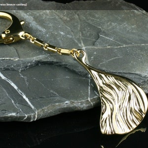 May include: A gold-colored key chain with a wave-shaped pendant. The chain is made of metal and has a lobster clasp. The pendant is made of bronze and has a textured surface.