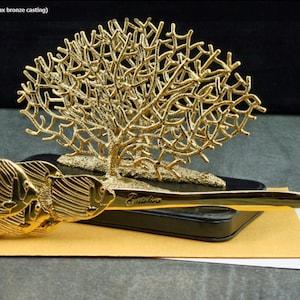 May include: Gold-toned letter holder featuring a sea fan design and a group of fish. The holder is made of metal and has a black base. The text "Galler" is visible on the base.