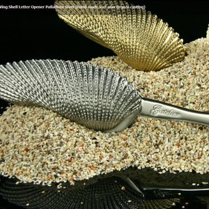 May include: A silver letter opener shaped like a seashell with the word "Gautier" engraved on the handle. The opener is resting on a bed of white sand with two seashells in the background.
