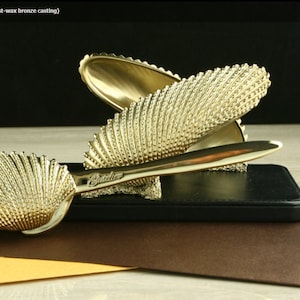 May include: A gold-colored metal letter holder shaped like a seashell with a textured surface. The holder is made of titanium and has a lost-wax bronze casting.