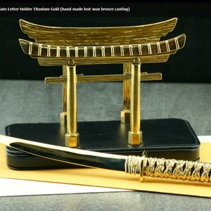 May include: A gold-colored letter opener with a decorative handle in the shape of a Japanese sword. The opener is resting on a black base with a gold-colored Japanese gate sculpture on top.