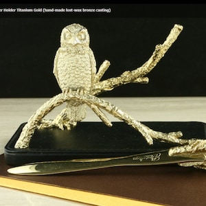 May include: A gold owl letter opener with a detailed design. The owl is perched on a branch and is holding a letter opener in its beak. The letter opener is made of titanium gold and is hand-made using the lost-wax bronze casting method.