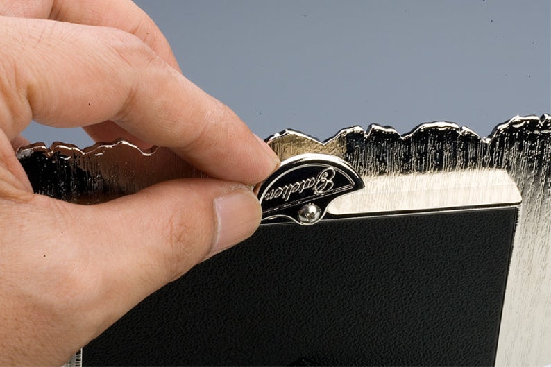 May include: A silver-toned picture frame with a decorative, scalloped edge. The frame has a black backing and a small, silver-toned latch with the word "Barclay" engraved on it.