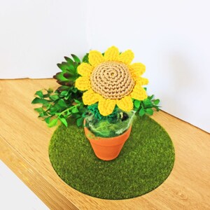 Könnte beinhalten: Eine gehäkelte Sonnenblume in einem Terrakotta-Topf, der auf einer grünen Grasmatte steht. Die Sonnenblume hat gelbe Blütenblätter und eine braune Mitte.