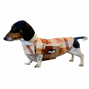 May include: A brown and tan plaid dog coat with a patch that says "Dachsja Jackets". A dachshund dog is wearing the coat.