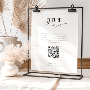 May include: A black metal sign holder displaying a white paper with the text "FUTURE Fund pot". The paper includes a QR code and text about a future fund. Dried flowers and decorative elements are in the background.