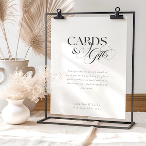 Può includere: Un porta cartello in metallo nero che mostra una carta bianca con le parole "CARDS & Gifts" in elegante scrittura. La carta include un messaggio sulla generosità, con una data e dei nomi in basso. Fiori secchi e pampas grass decorano lo sfondo.