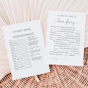 May include: Two white paper cards with fill-in-the-blank prompts for a wedding or bridal shower. One card says "Start Here" and the other says "Olivia & Luke's Love Story". The cards are on a woven surface.
