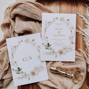 Pode incluir: Dois cartões de convite de casamento com desenhos de grinaldas florais. Um cartão exibe as iniciais "G|A", o outro diz "Grace e Antonio" com detalhes do evento. Uma cera de vedação e um carimbo dourados também estão presentes.