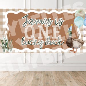May include: A banner with the text "James is ONE lucky duck" in a playful font. The banner has a brown and white checkered background with a duck wearing a party hat and balloons. Decorative grass is on the left.