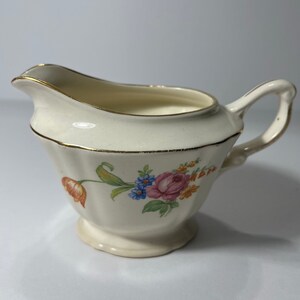May include: A small, white creamer with a gold rim and floral design. The creamer has a delicate, feminine look and is perfect for adding a touch of elegance to any table setting.