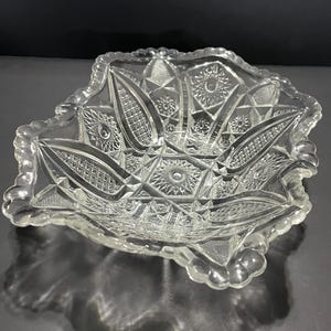May include: Clear glass serving bowl with a detailed, geometric pattern. The bowl has a scalloped edge with a beaded border. The interior features starburst and diamond designs. This decorative bowl is ideal for serving snacks or displaying decorative items.