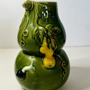 May include: A gourd-shaped ceramic vase with a glossy dark green glaze and a crackle finish. The surface features raised relief details including green leaves, curling vines, a small yellow gourd, and a small white and black snail-like figure.