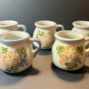 May include: Five ceramic mugs with floral designs. The mugs are off-white with a light blue rim and handle. The floral pattern includes peach, yellow, green, and blue colors. The mugs are arranged on a dark surface.