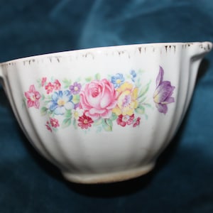 May include: A white ceramic bowl with a floral design. The bowl has a scalloped rim and two handles. The flowers are pink, yellow, blue, and purple.