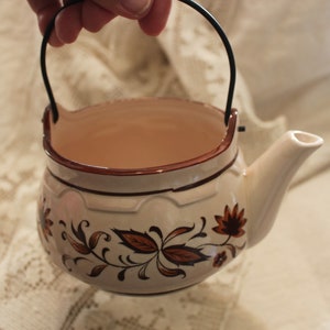 May include: A white ceramic teapot with a brown floral design and a black wire handle. The teapot has a spout and a rounded body.