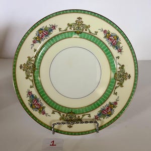 May include: A vintage porcelain plate with a green and gold floral design. The plate has a cream-colored background and a white center.