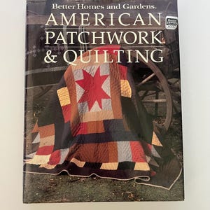 May include: A book titled "American Patchwork & Quilting" by Better Homes and Gardens. The cover features a colorful quilt with a red star design. The quilt is draped over a wooden wagon wheel.