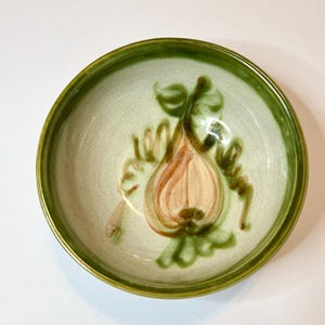 May include: A small, round ceramic bowl with a light green interior and a darker green rim. The bowl features a hand-painted design of a pear with green leaves and vines in shades of brown and green. The bowl is on a white background.
